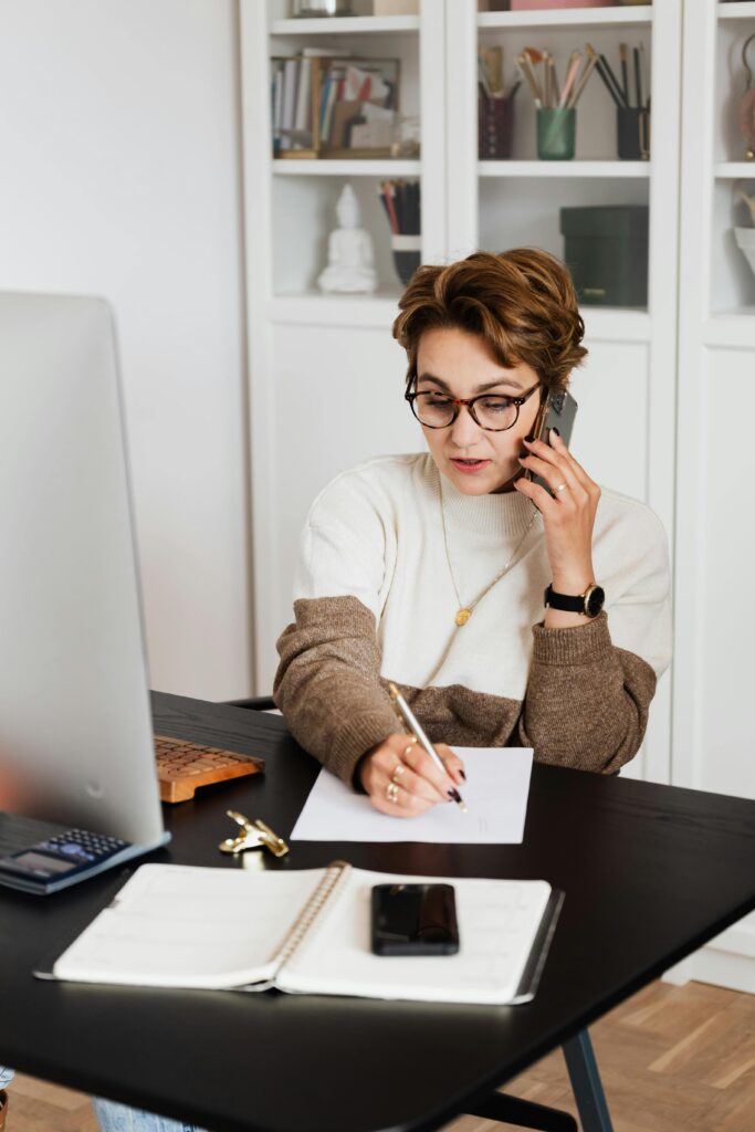 A woman working remotely from home, engaging in a phone call while taking notes.